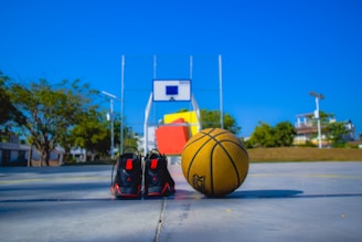 A basketball and a pair of black sports shoes are placed on an outdoor basketball court. A basketball hoop with a backboard is visible in the background, and the court is surrounded by trees and some buildings under a clear blue sky.
