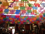 Colorful papel picado banners hang from the ceiling, casting a vibrant display of intricate patterns in red, blue, purple, green, yellow, and orange. The room below is dimly lit but decorated with textiles and various crafts, suggesting a festive or cultural ambiance.