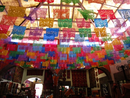 Colorful papel picado banners hang from the ceiling, casting a vibrant display of intricate patterns in red, blue, purple, green, yellow, and orange. The room below is dimly lit but decorated with textiles and various crafts, suggesting a festive or cultural ambiance.