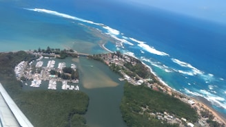 An aerial view of a coastal landscape with a marina filled with boats, surrounded by lush greenery and a vibrant blue ocean with waves crashing against the shoreline. A small town can also be seen near the coast along with sandy beaches.