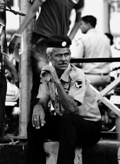 An older man in a uniform, seated pensively on a low ledge. He wears a hat and appears to be part of a security or law enforcement team, as suggested by the badges on his shirt. The setting seems urban, with other people walking past in the background, slightly blurred, indicating motion and a busy environment.