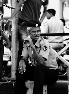 An older man in a uniform, seated pensively on a low ledge. He wears a hat and appears to be part of a security or law enforcement team, as suggested by the badges on his shirt. The setting seems urban, with other people walking past in the background, slightly blurred, indicating motion and a busy environment.
