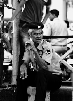 An older man in a uniform, seated pensively on a low ledge. He wears a hat and appears to be part of a security or law enforcement team, as suggested by the badges on his shirt. The setting seems urban, with other people walking past in the background, slightly blurred, indicating motion and a busy environment.