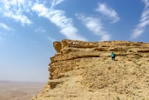 A rugged cliffside under a bright blue sky with scattered clouds. A few people are seen climbing the rocky terrain, suggesting an adventurous outdoor activity. The landscape extends into a vast desert, adding to the sense of openness and isolation.