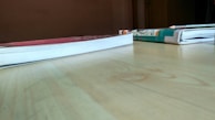 Two books lie flat on a wooden table, one with a red cover and the other with a colorful illustrated cover. A pencil is placed on top of the book with the illustrated cover. The table surface is light brown, and the background has a plain wall.