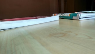Two books lie flat on a wooden table, one with a red cover and the other with a colorful illustrated cover. A pencil is placed on top of the book with the illustrated cover. The table surface is light brown, and the background has a plain wall.