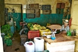 A small, rustic shop featuring piles of bananas, plastic crates, and a scale on a counter. A man in a hat sits beside some bottles, with woven baskets and a bicycle visible in the background. The walls are aged, with peeling paint and storage shelves.