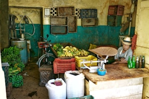 A small, rustic shop featuring piles of bananas, plastic crates, and a scale on a counter. A man in a hat sits beside some bottles, with woven baskets and a bicycle visible in the background. The walls are aged, with peeling paint and storage shelves.