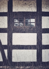 A section of a traditional timber-framed structure with dark wooden beams contrasting against white plaster. The central feature is a small, rectangular window divided into four panes, through which some foliage can be seen. The plaster exhibits a textured, rustic appearance, adding to the historical aesthetic of the building.