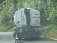 A person is hanging onto the back of a large fuel truck on a paved road surrounded by greenery. The truck has a warning symbol for flammable materials. Another person on a bicycle is riding close to the back of the truck. The scene appears to be in a rural or forested area.