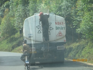 A person is hanging onto the back of a large fuel truck on a paved road surrounded by greenery. The truck has a warning symbol for flammable materials. Another person on a bicycle is riding close to the back of the truck. The scene appears to be in a rural or forested area.