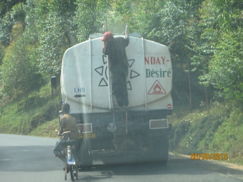 A person is hanging onto the back of a large fuel truck on a paved road surrounded by greenery. The truck has a warning symbol for flammable materials. Another person on a bicycle is riding close to the back of the truck. The scene appears to be in a rural or forested area.