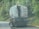 A person is hanging onto the back of a large fuel truck on a paved road surrounded by greenery. The truck has a warning symbol for flammable materials. Another person on a bicycle is riding close to the back of the truck. The scene appears to be in a rural or forested area.