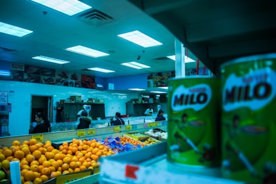Interior of a grocery store with various fresh produce and canned goods visible. Brightly lit ceiling illuminates oranges, potatoes, and large cans of Milo placed on shelves. Several people are seen shopping in the background near a seafood section.