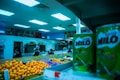 Interior of a grocery store with various fresh produce and canned goods visible. Brightly lit ceiling illuminates oranges, potatoes, and large cans of Milo placed on shelves. Several people are seen shopping in the background near a seafood section.