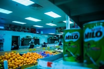 Interior of a grocery store with various fresh produce and canned goods visible. Brightly lit ceiling illuminates oranges, potatoes, and large cans of Milo placed on shelves. Several people are seen shopping in the background near a seafood section.