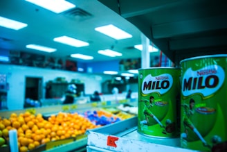 Shelves displaying large tins of Milo in the foreground of a grocery store, with vibrant lighting and colorful fruit like oranges in the background. The store setting includes visible shelves stocked with various items and a blue tinted ambiance from the overhead fluorescent lights.