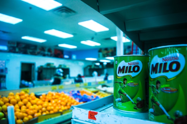 Shelves displaying large tins of Milo in the foreground of a grocery store, with vibrant lighting and colorful fruit like oranges in the background. The store setting includes visible shelves stocked with various items and a blue tinted ambiance from the overhead fluorescent lights.