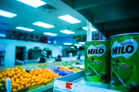 Shelves displaying large tins of Milo in the foreground of a grocery store, with vibrant lighting and colorful fruit like oranges in the background. The store setting includes visible shelves stocked with various items and a blue tinted ambiance from the overhead fluorescent lights.