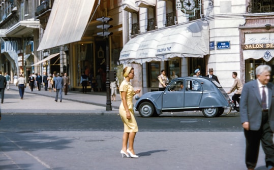 A busy city street scene features pedestrians walking by, classic cars parked alongside the road, and storefronts with awnings stretching out over the sidewalk. People are dressed in mid-20th century attire, and a woman in a yellow dress and white shoes walks across the road. The background includes a caf&eacute; and various shops.