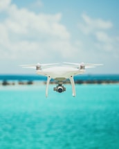 A drone is hovering in the foreground against a backdrop of a clear blue sky and a bright turquoise ocean. The design of the drone is sleek and modern, featuring a camera on its underside. The water and sky create a serene and expansive atmosphere.