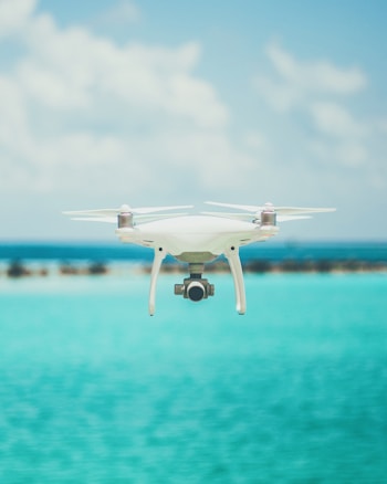 A drone is hovering in the foreground against a backdrop of a clear blue sky and a bright turquoise ocean. The design of the drone is sleek and modern, featuring a camera on its underside. The water and sky create a serene and expansive atmosphere.