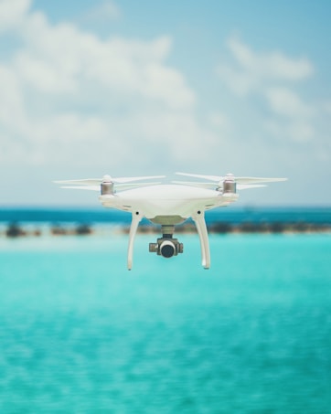 A drone is hovering in the foreground against a backdrop of a clear blue sky and a bright turquoise ocean. The design of the drone is sleek and modern, featuring a camera on its underside. The water and sky create a serene and expansive atmosphere.