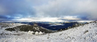 A snowy landscape with a group of white yurts nestled among hills. The foreground is covered in snow, while the background reveals distant mountains under a dramatic sky with patches of clouds. The lighting suggests a cool, possibly early morning ambiance.