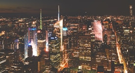A vibrant cityscape at night with numerous skyscrapers illuminated by bright lights. The skyline features a blend of modern architecture, highlighted by colorful advertisements and light displays. Streets are lined with glowing lines of traffic, exuding an energetic urban atmosphere.