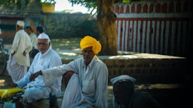 A group of men dressed in traditional white clothing are gathered outdoors. One man in the foreground is wearing a bright yellow turban, contrasting with the rest in white. They are seated or standing near a brick structure with a tree providing shade. The background includes a striped wall and more people in similar attire.