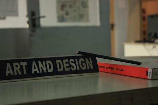 A close-up view of a table displaying a book titled 'Materials for Design' by Chris Lefteri. A black pencil rests on top of the book. In the foreground, there is a sign with the words 'Art and Design'. The background appears to be an indoor setting, slightly out of focus, with cool, muted lighting.