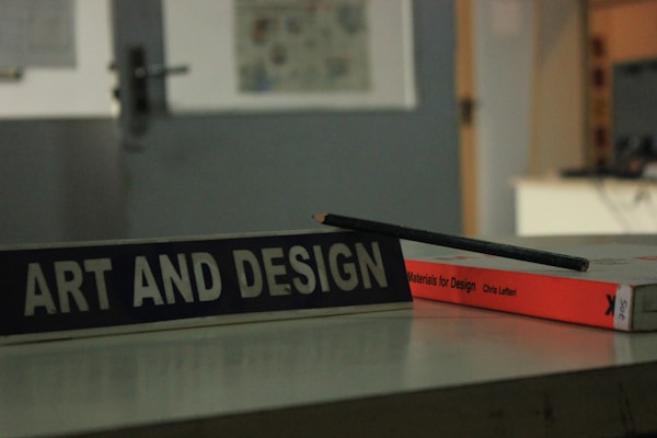 A close-up view of a table displaying a book titled 'Materials for Design' by Chris Lefteri. A black pencil rests on top of the book. In the foreground, there is a sign with the words 'Art and Design'. The background appears to be an indoor setting, slightly out of focus, with cool, muted lighting.