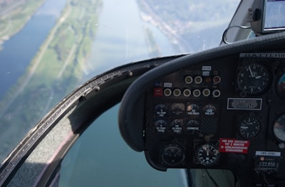 A close-up view of an aircraft cockpit showing various dials, gauges, and switches. The cockpit window reveals an aerial view of a river and landscape below, suggesting the aircraft is in flight.
