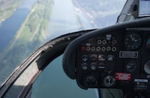 A close-up view of an aircraft cockpit showing various dials, gauges, and switches. The cockpit window reveals an aerial view of a river and landscape below, suggesting the aircraft is in flight.