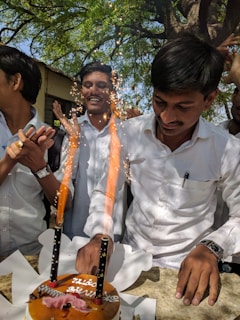 A group of people are gathered outdoors, celebrating with a cake that has lit sparklers on it. The scene is lively and joyful, with one person cutting the cake while others clap and cheer. The background features green trees and sunlight filtering through the leaves.