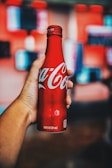 A hand holding a red Coca-Cola bottle with a blurred background that features soft, colorful shapes. The bottle is metallic and branded with the iconic white Coca-Cola script.