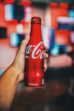 A hand holding a red Coca-Cola bottle with a blurred background that features soft, colorful shapes. The bottle is metallic and branded with the iconic white Coca-Cola script.
