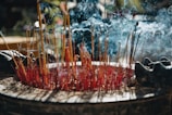 A large assortment of burning incense sticks are placed in a circular container, with smoke gently swirling in the air. The incense sticks appear red and yellow, surrounded by ash and half-burnt sticks. Subtle lighting creates an atmospheric effect, with hints of nature visible in the blurred background.