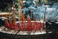 A large assortment of burning incense sticks are placed in a circular container, with smoke gently swirling in the air. The incense sticks appear red and yellow, surrounded by ash and half-burnt sticks. Subtle lighting creates an atmospheric effect, with hints of nature visible in the blurred background.