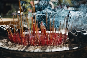 A large assortment of burning incense sticks are placed in a circular container, with smoke gently swirling in the air. The incense sticks appear red and yellow, surrounded by ash and half-burnt sticks. Subtle lighting creates an atmospheric effect, with hints of nature visible in the blurred background.