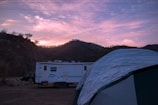 A serene campsite in a valley as dusk approaches. A large white recreational vehicle is parked next to a tent, with a background of rolling hills and a pink and purple hued sky.