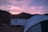 A serene campsite in a valley as dusk approaches. A large white recreational vehicle is parked next to a tent, with a background of rolling hills and a pink and purple hued sky.
