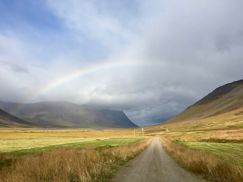 caminho de terra entre o campo marrom e verde que leva a um arco-íris sob o céu nublado azul e branco