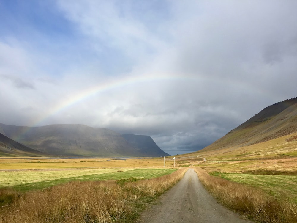 dirt path between brown and green field leading to a rainbow under blue and white cloudy sky