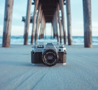 black and gray Canon AE-1 camera on gray sand under brown dock near body of water at daytime