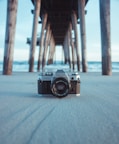 black and gray Canon AE-1 camera on gray sand under brown dock near body of water at daytime