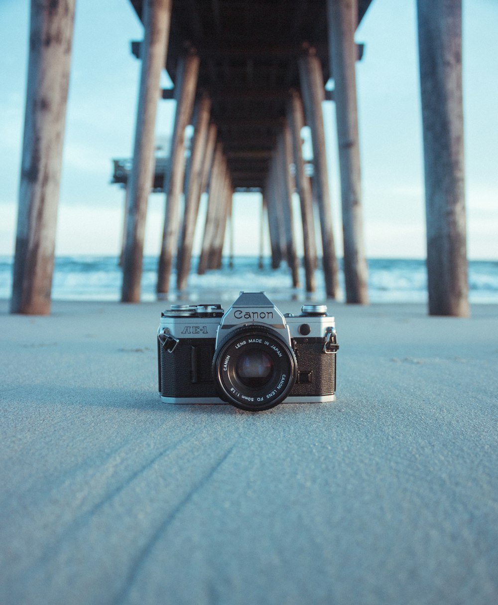 câmera Canon AE-1 preta e cinza na areia cinza sob a doca marrom perto do corpo de água durante o dia