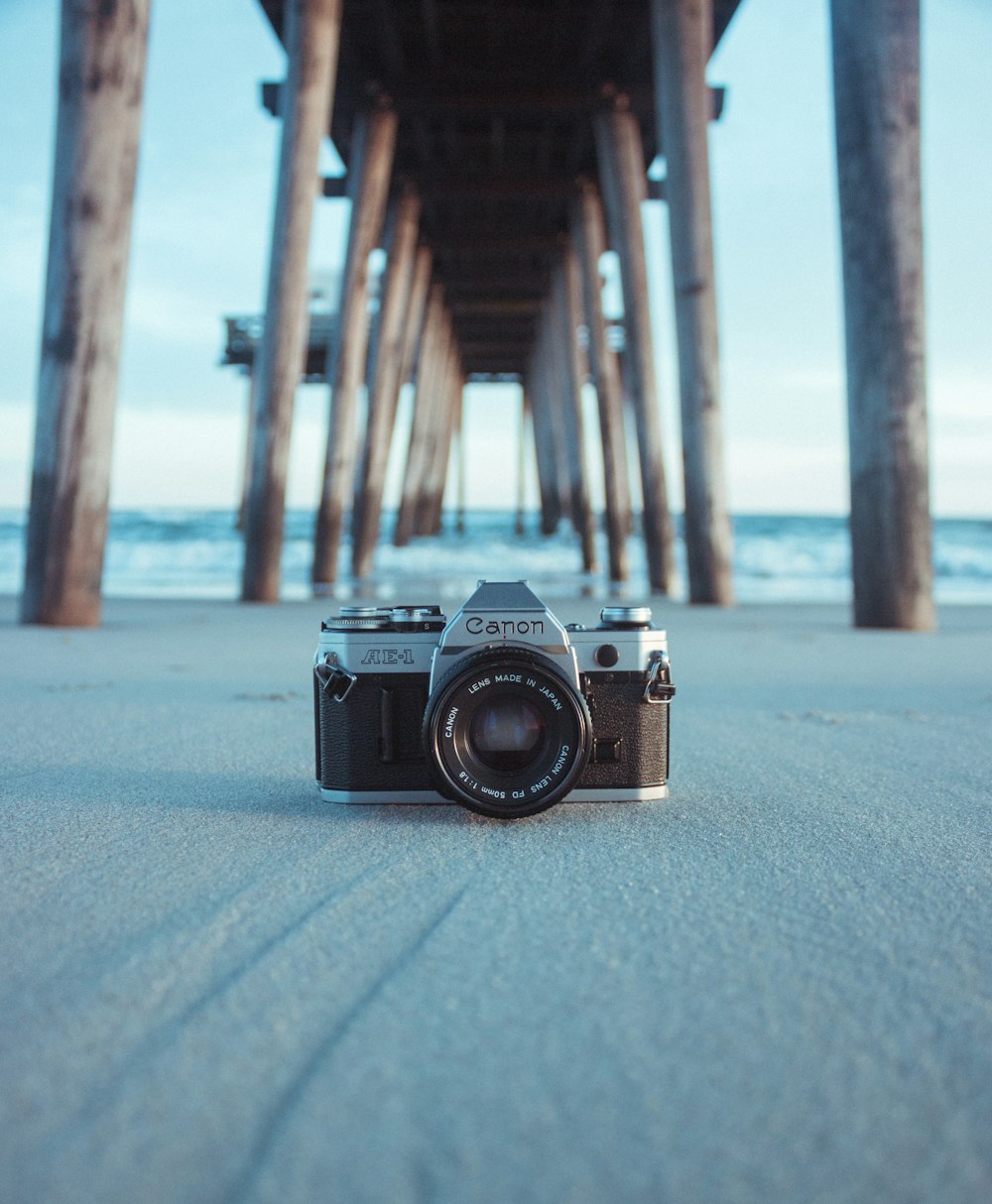 Cámara Canon AE-1 negra y gris en arena gris debajo de un muelle marrón cerca del cuerpo de agua durante el día