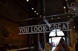 A person holding a camera is taking a photo in what appears to be an industrial-style interior with exposed brick walls and metal fixtures. There is a large sign reading 'YOU LOOK GREAT' prominently displayed in bold white letters. The environment is dimly lit with spotlighting from the ceiling, and there is a large arched window in the background letting in natural light.