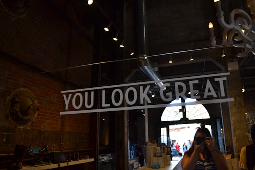 A person holding a camera is taking a photo in what appears to be an industrial-style interior with exposed brick walls and metal fixtures. There is a large sign reading 'YOU LOOK GREAT' prominently displayed in bold white letters. The environment is dimly lit with spotlighting from the ceiling, and there is a large arched window in the background letting in natural light.